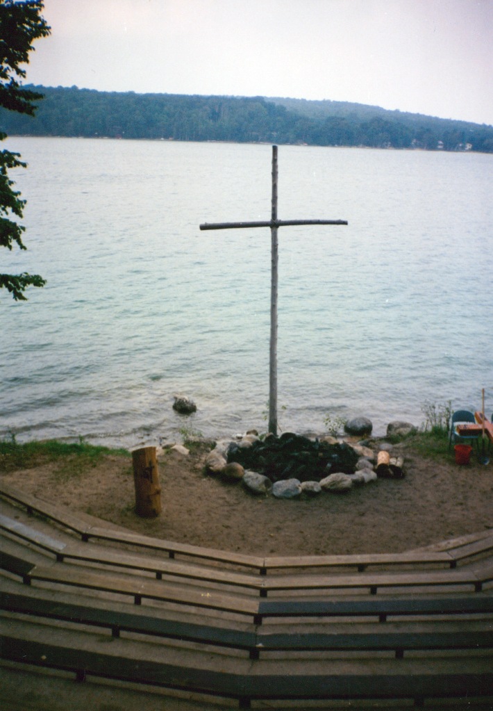 A wood cross in front of a small lake with trees on the other side of the lake. In front of the cross is a fire bowl surrounded by large stones. There is a tree trunk podium to the left of the fire bowl. Out surrounding the fire bowl and beach area are rounded rows of wood benches going uphill (although I guess you can't quite tell they go up hill from the photo).